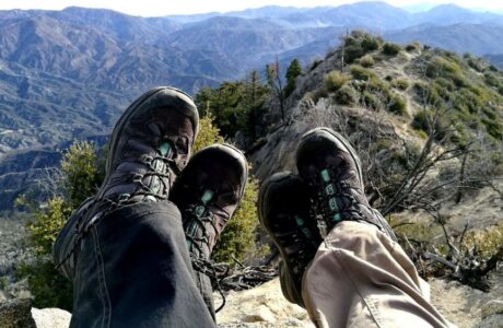 dwie osoby w czarnych butach trekkingowych leżące na skraju zbocza z widokiem na góry