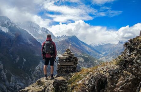 mężczyzna w czerwonej kurtce, czarnym plecaku i czarnych krótkich spodenkach stojący na skraju góry patrzący na góry